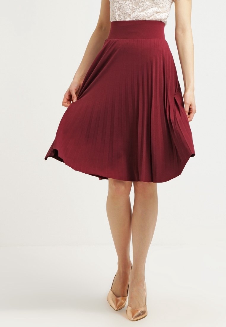 Donna che indossa una gonna midi plissettata rossa e tacchi beige a punta, che tiene i bordi della gonna, in piedi contro uno sfondo chiaro e uniforme.