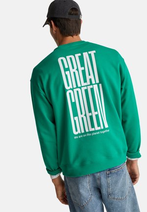 Uomo che indossa un berretto nero e una felpa verde con grande testo bianco "GREAT GREEN" e un testo più piccolo sotto.