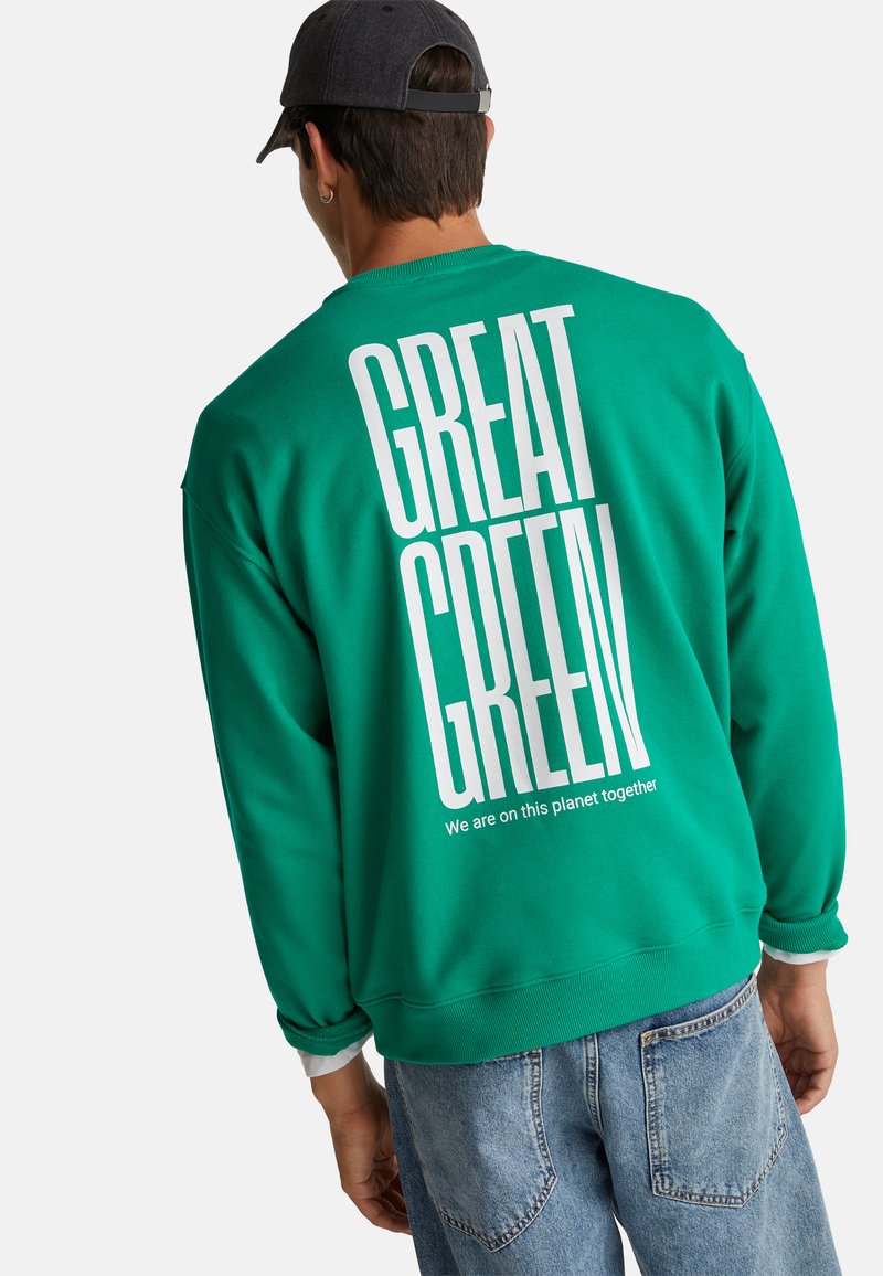 Homme portant une casquette noire et un sweat-shirt vert avec un grand texte blanc "GREAT GREEN" et un texte plus petit en dessous.