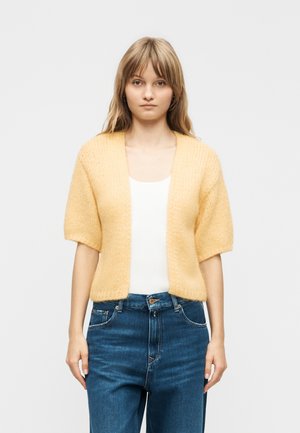 Jeune femme portant un cardigan en tricot à manches courtes jaune par-dessus un haut blanc et un jean bleu taille haute, debout devant un fond uni.