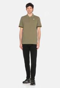 Polo shirt verde oliva con polsini a righe gialle e nere, abbinato a jeans neri e sneaker scuri. Design semplice e aderente, maniche corte.