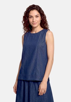 Donna con capelli ricci che indossa un top di jeans blu senza maniche e una gonna abbinata, in piedi davanti a uno sfondo semplice.