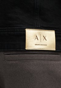 Μαύρο παντελόνι από τζιν με υφασμάτινη χρυσή ετικέτα που φέρει το λογότυπο "A|X" και την επιγραφή "ARMANI EXCHANGE" χαραγμένη από κάτω, τοποθετημένη πάνω σε απαλό γκρίζο ζωνάρι μέσης.