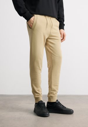 Bărbat purtând pantaloni jogging bej, adidași negri și un top cu mâneci lungi negre, stând cu o mână în buzunar pe un podea gri.