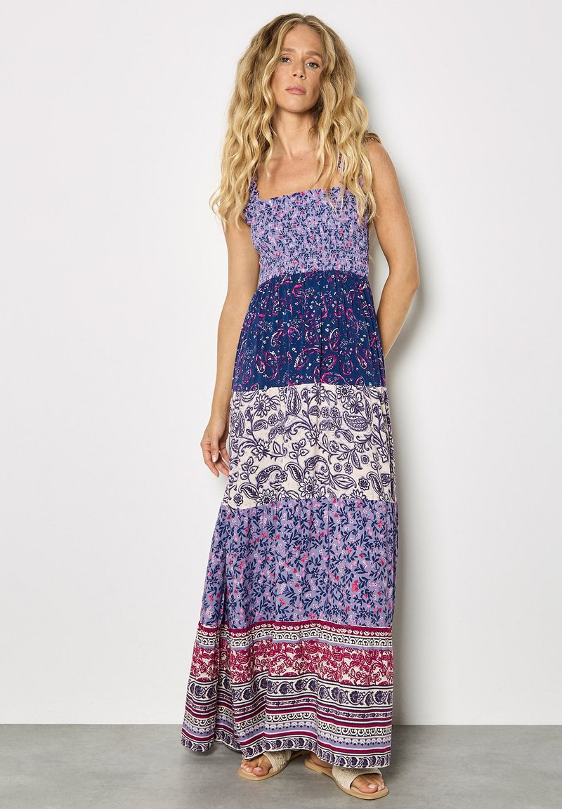 Femme aux longs cheveux ondulés portant une robe maxi à volants multicolore avec des motifs floraux et cachemire, debout contre un mur blanc.