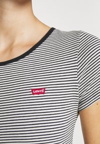 Tricou cu dungi alb-negru, cu guler rotund și un mic patch roșu cu logo-ul Levi's pe partea stângă a pieptului.