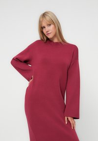 Abito lungo bordeaux a maniche lunghe con collo alto, vestibilità comoda e texture morbida. Il design include maniche ampie e tasche laterali.
