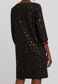 Robe noire avec des motifs de feuilles métalliques, manches trois-quarts et un détail de dos en forme de trou de serrure. Texture lisse et coupe droite.