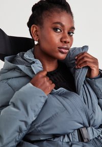 Pilkas paminkštintas švarkas su aukšta apykakle, užtrauktuku ir diržu. Lygus paviršius ir dideli siūlės. Dėvimas ant juodos bluzonos.