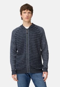 Gebreid vest met rits in donkerblauwe tinten, met gehaakte patronen, geribbelde manchetten en een ronde halslijn. Draagt over een wit T-shirt.