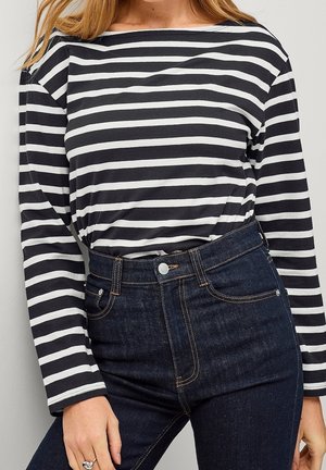 Kobieta w czarno-białej, poziomo paskowanej bluzce z długim rękawem, wpuszczonej do ciemnych, wysokich dżinsów z kontrastowym przeszyciem.