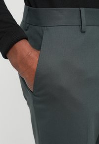 Παντελόνια σε σκουρόγρινο ύφασμα με υφή, διαθέτουν πλαϊνή τσέπη και θηλιά για ζώνη. Κοντινή όψη του υφάσματος και του χεριού στην τσέπη.