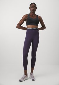 Sujetador deportivo negro combinado con leggins morados que cuentan con una cinturilla negra de Nike Pro, usados con zapatillas deportivas de color morado claro. Textura de tela suave.