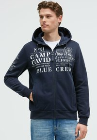 Sudadera con cremallera de color navy con capucha forrada, estampados de texto gráfico en blanco, bolsillos frontales, puños y dobladillo acanalados. Diseño casual adecuado para superponer.