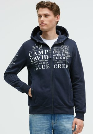 Navy zip-up hoodie with a lined hood, graphic text prints in white, front pockets, ribbed cuffs, and hem. Casual design suitable for layering.