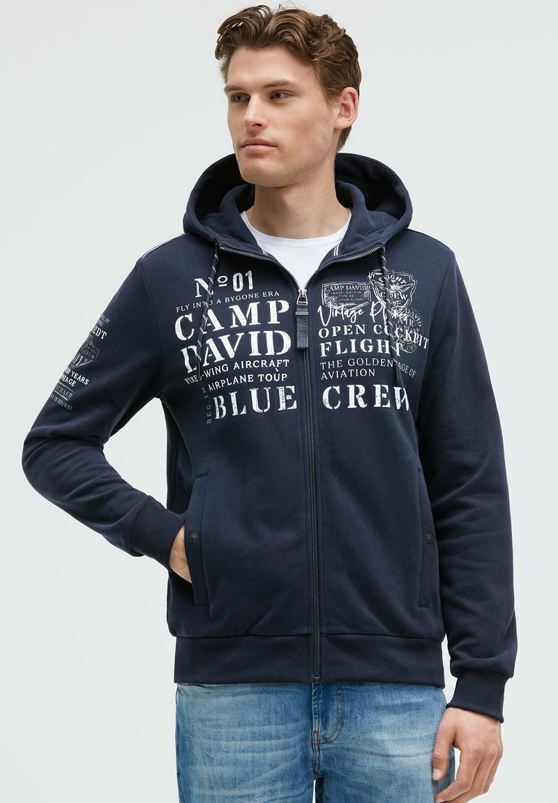 Sudadera con cremallera de color navy con capucha forrada, estampados de texto gráfico en blanco, bolsillos frontales, puños y dobladillo acanalados. Diseño casual adecuado para superponer.