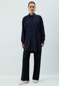 Navyblå ternet skjorte med lange ærmer, kombineret med sorte bukser med brede ben og hvide sneakers, sat mod en lys baggrund.