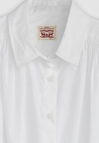 Vit bomullsskjorta med knapp-down krage, texturerat mönster och en etikett med röda grafik som visar Levi Strauss & Co. och storlek S.