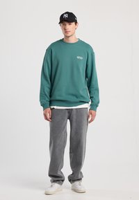 Teal sweatshirt with ribbed cuffs and hem, featuring a small NYC logo. Paired with loose gray jeans and white sneakers. Black cap.
