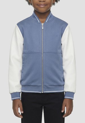 Enfant portant une veste zippée bleue et blanche avec un pantalon noir, debout devant un fond gris uni.