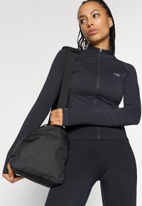 Melnas sporta jakas ar rāvējslēdzēju, izgatavota no stiepjoša auduma, kombinēta ar melniem legingiem un strukturēta auduma sporta somu. Garajām piedurknēm ir thumb hole (pirksta caurumi).