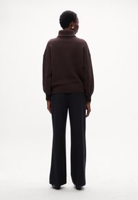 Maglione a collo alto in maglia grossa marrone con spalle scese, abbinato a pantaloni a gamba larga a righe sottili a vita alta, visti da dietro.