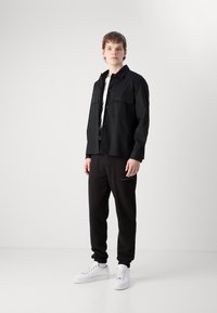 Zwarte button-up jas met twee borstzakken, gecombineerd met zwarte sweatpants met logo en witte sneakers. Eenvoudig, modern ontwerp.