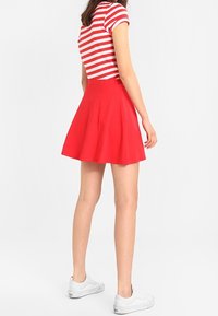 Roter ausgestellter Rock, glatte Textur, hohe Taille. Kombiniert mit einem rot-weiß gestreiften Kurzarmoberteil und getragen mit weißen Sneakers.
