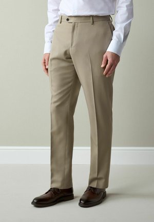 Mann trägt beige Anzughose, weißes Hemd mit Knöpfen und dunkelbraune Anzugsschuhe, steht vor einem schlichten hellen Hintergrund.