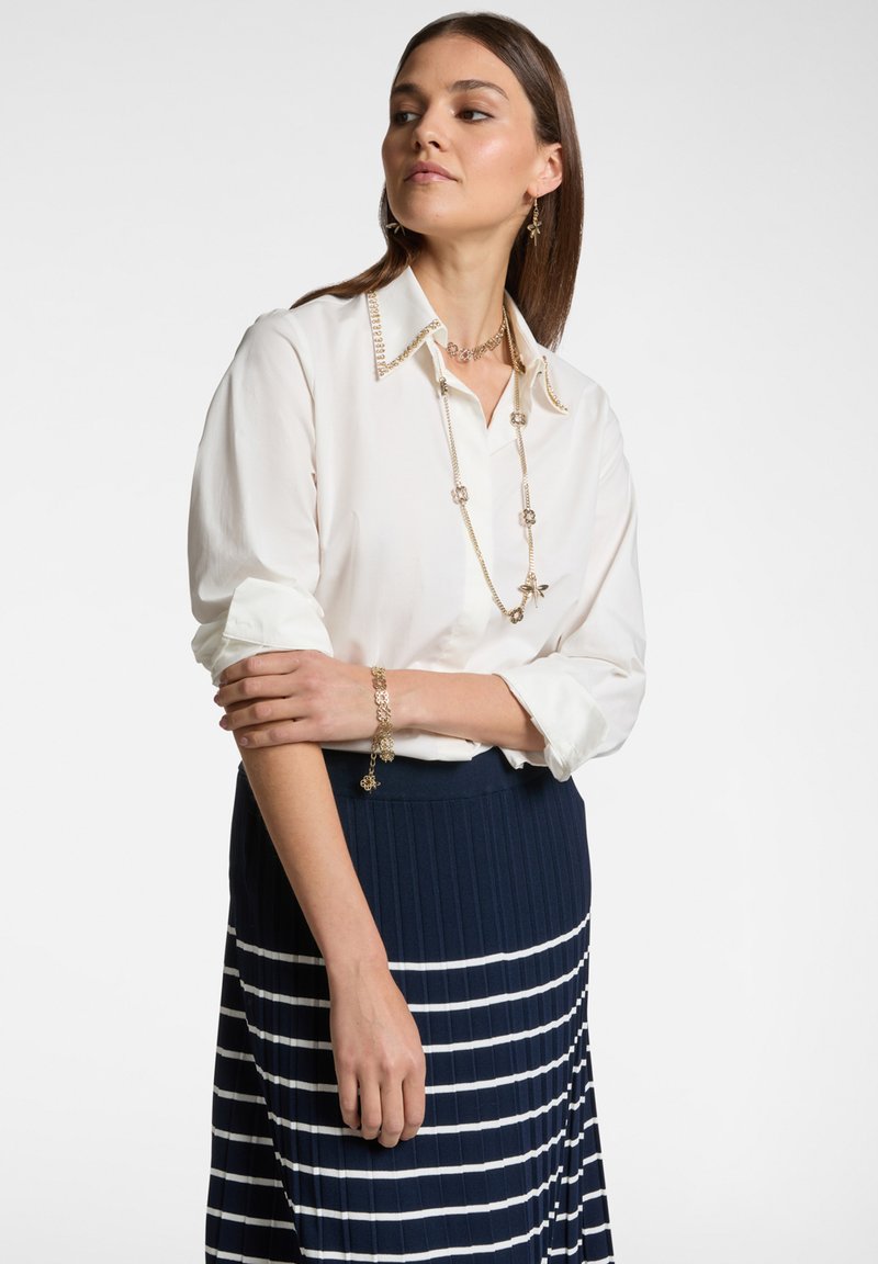 Blusa bianca con dettaglio collo decorativo, abbinata a una gonna plissettata blu navy con strisce bianche orizzontali. Gli accessori includono collane a più strati e un braccialetto.