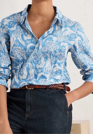 Niebieska koszula na guziki z białym kwiatowym wzorem, wpuszczona do ciemnych dżinsów z brązowym plecionym paskiem, z jedną ręką w kieszeni.