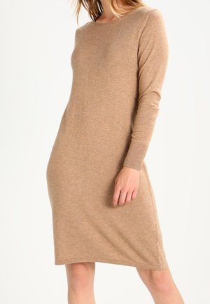 Femme portant une robe beige en maille à manches longues, longueur genou, debout devant un fond blanc uni.