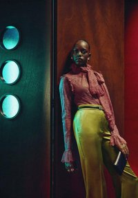 Blusa de encaje en rosa claro con mangas con volantes y detalle de lazo, combinada con pantalones amarillos brillantes y un bolso negro. Fondo de madera con acentos verdes.