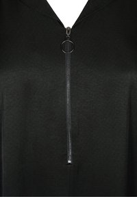 Indumento in tessuto nero con una cerniera frontale dotata di una linguetta circolare. Il tessuto appare liscio con una leggera lucentezza e un design con cappuccio.