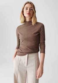 Brauner, enganliegender Rollkragenpullover mit subtiler metallischer Ausstrahlung und Dreiviertelärmeln, kombiniert mit hellbeigen, maßgeschneiderten Hosen.