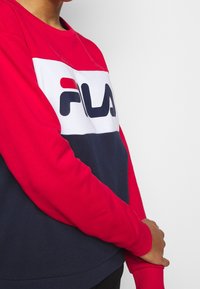 Persoon die een rode, witte en marineblauwe FILA-trui draagt, staand met één hand die de tegenovergestelde pols vasthoudt tegen een effen lichte achtergrond.