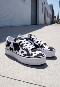 Paire de baskets basses avec motif vache noir et blanc et lacets blancs sur un trottoir en béton ensoleillé à côté d’un bâtiment en briques blanches.