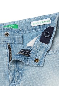 Pantalones de mezclilla azul claro con cremallera, cierre de botón y tejido texturizado. La etiqueta dice "BRUCE-STRAIGHT". Incluye acentos en azul marino contrastantes.
