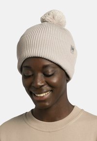 Beige knitted beanie with a ribbed texture and a pom-pom on top. Features a small logo patch near the brim. Worn by a person smiling.