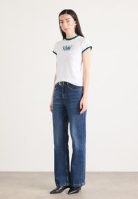 White short-sleeve t-shirt with green trim and logo, paired with high-waisted blue denim jeans and black pointed-toe heels.