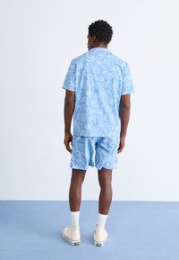 Lys blå kortermet skjorte og matchende shorts med hvit blomsterdesign. Laget av bomull, avslappet passform, kombinert med kremfargede sneakers og hvite sokker.
