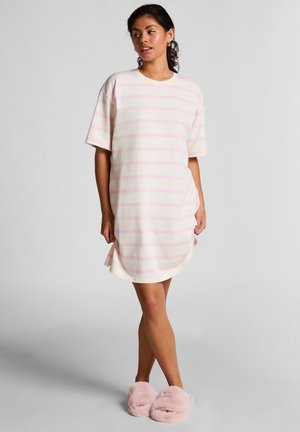 Woman standing in light cream and pink striped short-sleeve dress and pink fuzzy slippers against plain gray background.