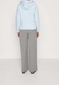 Persona de pie, de espaldas, vistiendo una sudadera ligera azul, pantalones gris de pierna ancha y zapatos oscuros, contra un fondo blanco liso.