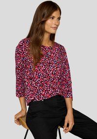 Frau mit langen braunen Haaren, die einen roten, lila und schwarz gemusterten Langarmpullover und schwarze Hosen trägt, sitzt auf einem hölzernen Hocker.