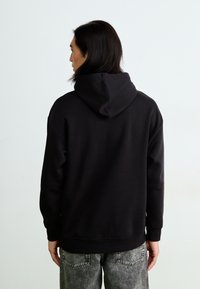 Sort hoodie med kængurulomme og justerbar snøre. Har ribbede manchetter og kant. Båret med grå slidte jeans. Bageste udsigt.