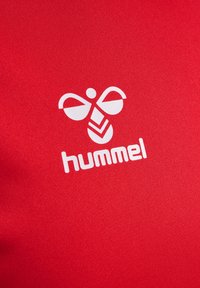 Wit Hummel-logo met gestileerde bij-vorm en tekst op een rode stoffen achtergrond.