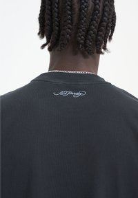 Černé bavlněné tričko s kulatým výstřihem, na zadní straně poblíž výstřihu má bílé vyšívané logo "Ed Hardy". Hladký materiál.