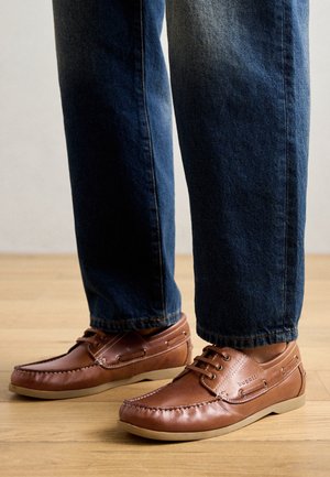 Zapatos mocasines marrones de cuero con cordones usados con vaqueros azul oscuro en un suelo de madera clara.