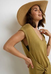 Abito verde oliva a maniche corte con scollatura a V, dettaglio annodato in vita e lati aperti, accessorato con un cappello di paglia a tesa larga.
