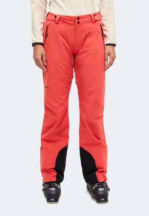 Personne portant un pantalon de ski rouge corail avec des poignets renforcés noirs et des chaussures de ski noires, debout, les mains détendues le long du corps.